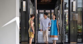 A smiling couple enters the exclusive Club InterContinental lounge at the luxury InterContinental Chiang Mai The Mae Ping hotel, greeted by a staff member in traditional Thai attire at the elegant black-framed glass entrance.